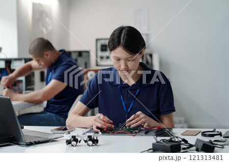 Skilled engineer soldering circuit board at modern tech lab, concentrating on intricate electronic components. Background features colleague working at another workstation Skilled engineer soldering circuit board at modern tech lab, concentrating on intricate electronic components. Background features colleague working at another workstation 121313481