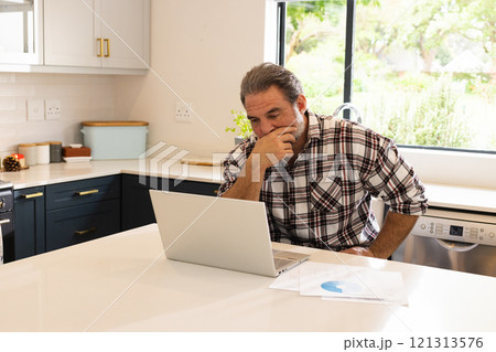 Mature man working on laptop in kitchen, focusing on online tasks, at home Mature man working on laptop in kitchen, focusing on online tasks, at home 121313576