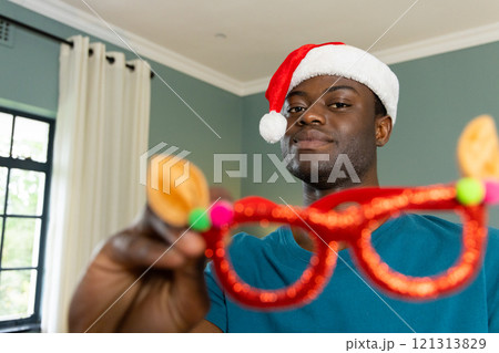 Christmas time, festive african american man wearing santa hat holding reindeer glasses at home 121313829