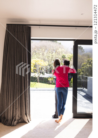 Happy diverse couple embracing and looking through window at home, copy space 121314472