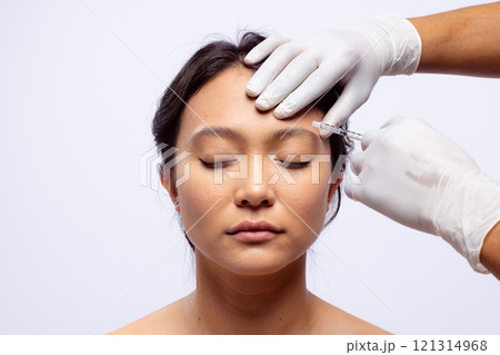Hands of beautician in white surgical gloves injecting botox in brow of asian woman, copy space 121314968