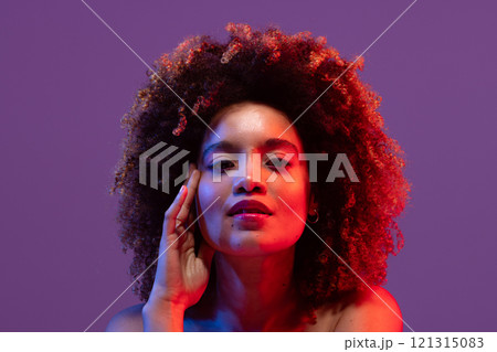 Biracial woman with curly hair, touching face in red light on purple background Biracial woman with curly hair, touching face in red light on purple background 121315083