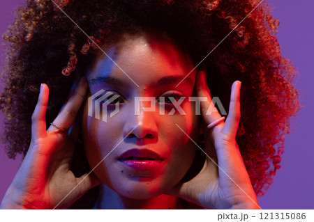 Biracial woman with curly hair, touching temples, in red and blue light with purple background 121315086