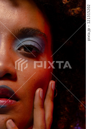 Half portrait of biracial woman with curly hair and blue eye shadow touching face with hand Half portrait of biracial woman with curly hair and blue eye shadow touching face with hand 121315202