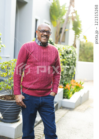 Portrait of happy senior african american man wearing glasses and red jumper standing outside house 121315268