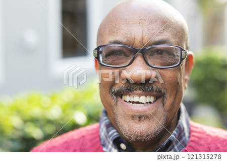 Happy senior african american man wearing glasses and red jumper standing outside house 121315278