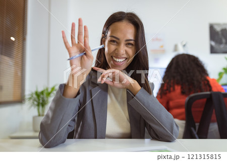 Smiling biracial businesswoman sitting at desk making video call waving in modern office 121315815