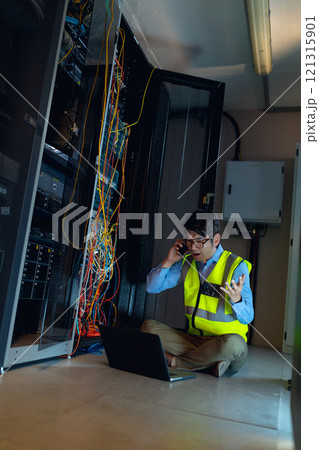 Asian male engineer with laptop talking on smartphone while inspecting in computer server room 121315901