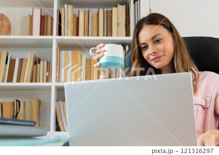 Woman working from home, holding coffee mug, focused on laptop screen 121316297