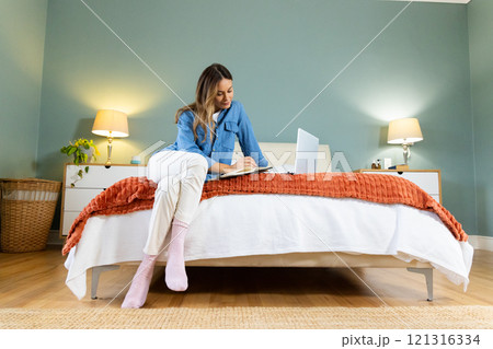 Woman sitting on bed writing in notebook with laptop, cozy bedroom setting, at home 121316334