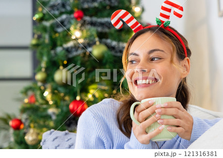 Smiling woman with festive headband enjoying hot drink by Christmas tree, at home Smiling woman with festive headband enjoying hot drink by Christmas tree, at home 121316381