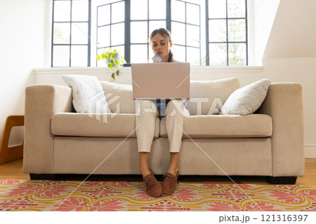 Woman working on laptop at home, sitting on cozy sofa, focused and thoughtful 121316397