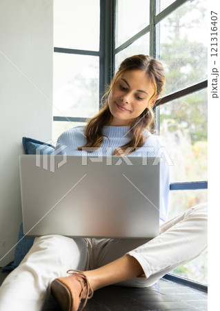 Smiling woman using laptop by window, enjoying cozy home atmosphere 121316407
