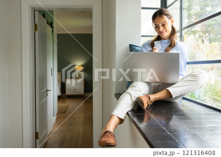 Woman sitting by window using laptop and holding credit card, shopping online, at home 121316408