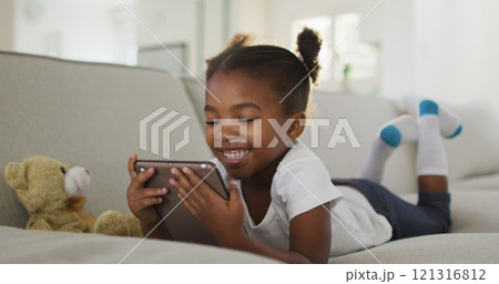 Happy african american girl lying on sofa using digital tablet at home 121316812
