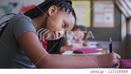 Biracial girl writing in a book while sitting on her desk at school 121316989