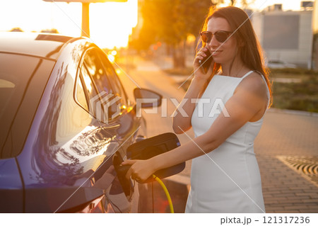 Woman charging electric car while talking on mobile phone during sunset in the street Woman charging electric car while talking on mobile phone during sunset in the street 121317236