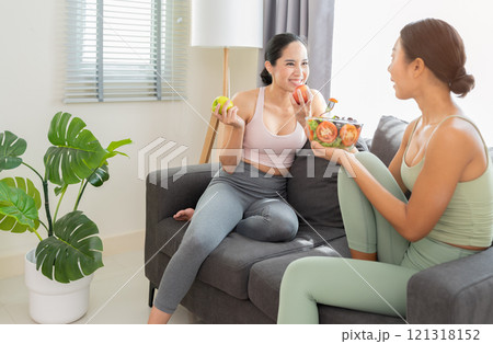 Two smiling women in active wear chatting and eating on couch in living room at home. One holding apple fruits while other holding salad bowl. Two smiling women in active wear chatting and eating on couch in living room at home. One holding apple fruits while other holding salad bowl. 121318152