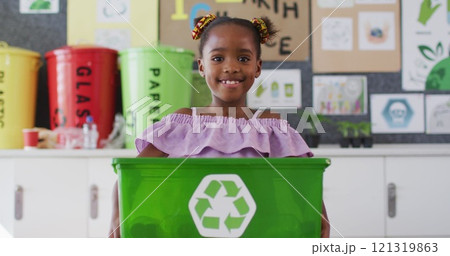 Happy african american schoolgirl standing in classroom, smiling, holding recycling bin Happy african american schoolgirl standing in classroom, smiling, holding recycling bin 121319863