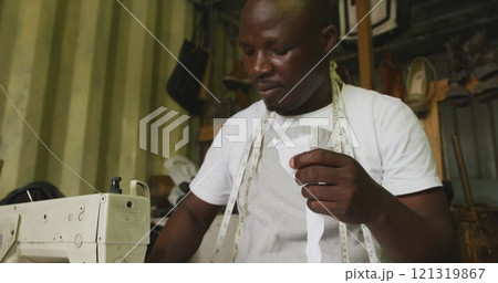 Front view of an African male tailor in a township workshop, sitting by a sewing machine and cutting 121319867