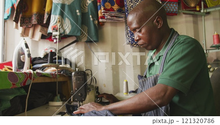 Side view of an African male tailor in a township workshop, using a sewing machine 121320786