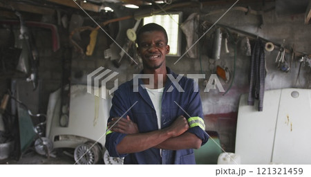 Portrait of a happy African male panel beater in a township workshop, looking at camera with arms cr 121321459
