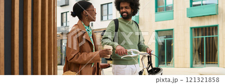 Two friends walking together while carrying coffee and a bicycle, having an engaging conversation in front of a modern building with wood and glass decor 121321858