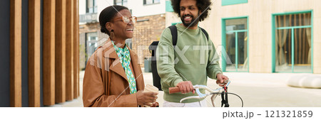 Two friends enjoying an outdoor walk, one holding a bicycle and other carrying a drink. Both are interacting and smiling while engaging in friendly banter 121321859