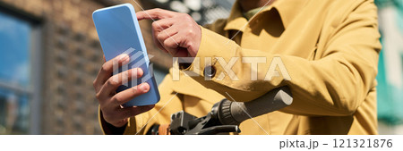 Person touching screen of a smartphone while resting hand on bicycle handle. Wearing a yellow jacket, the person appears to be outdoors, engaged in using the device 121321876