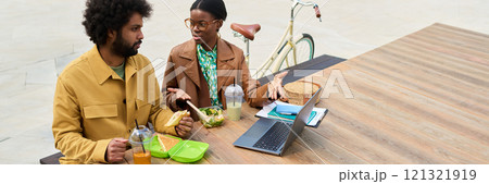 Two coworkers sitting at picnic table during lunch break outside, engaging in conversation about work projects while enjoying sandwiches and drinks 121321919