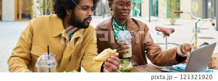 Two colleagues brainstorming by outside cafe table while enjoying brunch. Both focused on laptop screen and consuming iced coffee and salad during meeting Two colleagues brainstorming by outside cafe table while enjoying brunch. Both focused on laptop screen and consuming iced coffee and salad during meeting 121321920