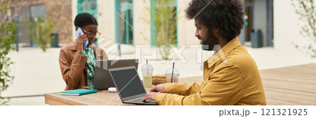 Colleagues working at outdoor table with laptops and beverages, engaging in conversation. One colleague multitasking while using a phone and wearing glasses 121321925