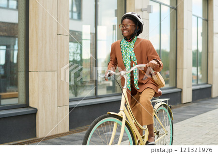 Woman riding bicycle along modern glass building while wearing casual outfit and helmet, smiling at something ahead, urban environment depicted Woman riding bicycle along modern glass building while wearing casual outfit and helmet, smiling at something ahead, urban environment depicted 121321976