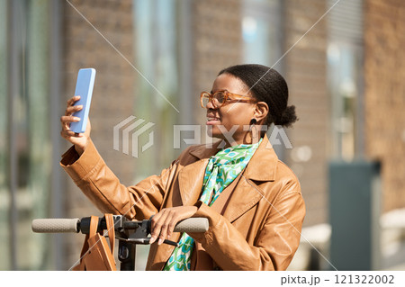 Young woman holding smartphone and smiling while standing next to bicycle handlebar in an urban setting on a sunny day 121322002