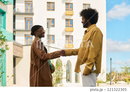 Two individuals engaging in handshake in outdoor urban setting with high-rise buildings in background. Both are smiling, conveying mutual respect and friendliness 121322305