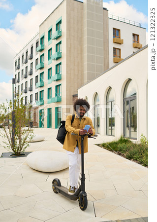 Individual standing on electric scooter while reading directions on tablet in urban area with modern architecture and greenery surrounding 121322325