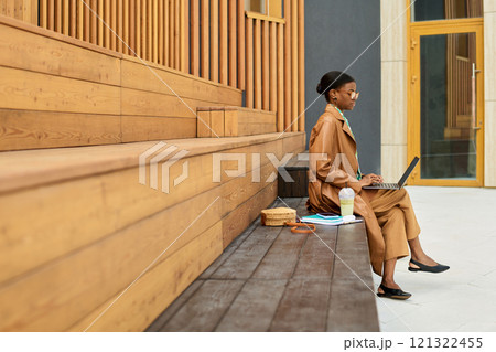 Woman sitting on wooden bench using laptop and drinking from coffee cup in an outdoor urban setting. Sunlight shining and creating shadows on wooden panels of the building behind her 121322455