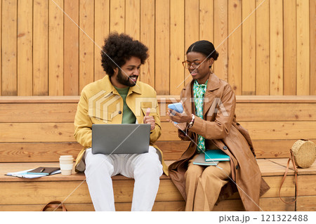 Two colleagues sitting on wooden bench sharing ideas using technology, with open laptop and digital tablet, showing collaborative working environment Two colleagues sitting on wooden bench sharing ideas using technology, with open laptop and digital tablet, showing collaborative working environment 121322488
