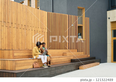 Two business people sitting on wooden steps outside modern building, talking and relaxing, background consists of contemporary architecture 121322489