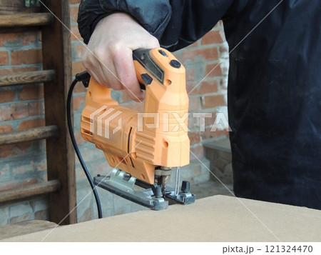 A carpenter's hand holds an orange jigsaw over a sheet of pressed cardboard or plywood to cut. Jigsaw work on wood in carpentry. 121324470