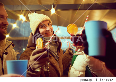 Young smiling man and woman in winter warm clothes cheerfully spending winter evening outdoor, drinking mulled wine, hot drinks. Young smiling man and woman in winter warm clothes cheerfully spending winter evening outdoor, drinking mulled wine, hot drinks. 121324542