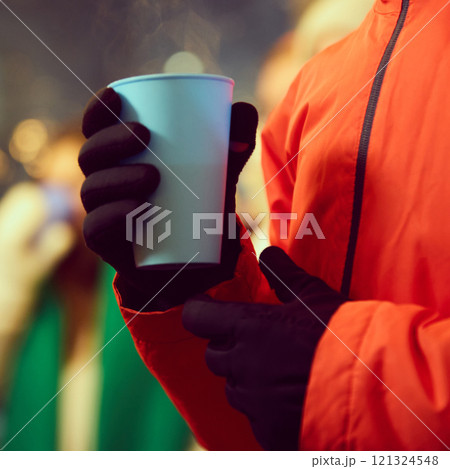 Close-up view of hand wearing black gloves holding blue cup with hot drink, set against blurred festive background. Outdoor fairs during holidays Close-up view of hand wearing black gloves holding blue cup with hot drink, set against blurred festive background. Outdoor fairs during holidays 121324548