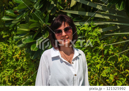 Woman in white shirt and sunglasses against dense green trees Woman in white shirt and sunglasses against dense green trees 121325992