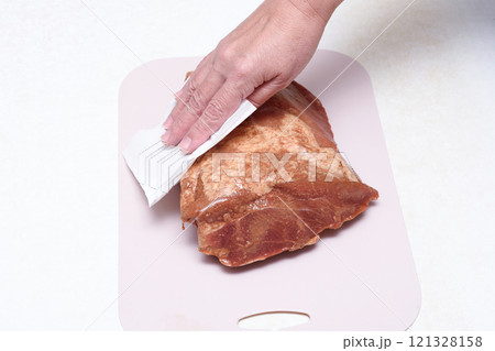 A woman's hand removes excess moisture from marinated meat using a paper napkin. 121328158