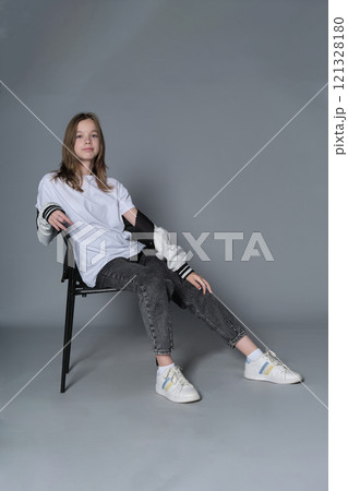 A teenage girl sits casually in a chair, her gaze steady and assured. Her posture reflects the self-confidence and ease of todays youth, embracing a minimalist style. 121328180