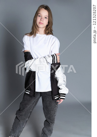 A young girl confidently adjusts her hair, wearing a varsity jacket over a casual outfit. This image captures the spirit of youth and self-expression, emphasizing individuality in today s modern world 121328207