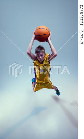 Aerial view of sportive, junior basketball athlete jumping in motion and throw ball into basket against grey studio background. Aerial view of sportive, junior basketball athlete jumping in motion and throw ball into basket against grey studio background. 121328572