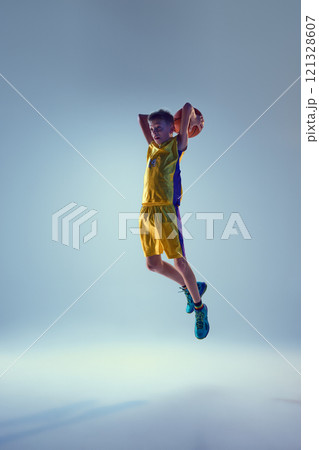 Little athletic boy, basketball player jumping in mid-air to get powerful slam dunk against grey studio background. Little athletic boy, basketball player jumping in mid-air to get powerful slam dunk against grey studio background. 121328607