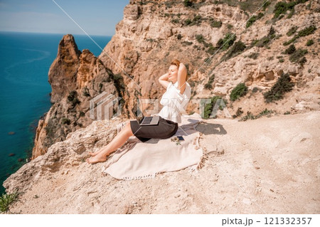 A woman is sitting on a blanket on a rocky beach, looking out at the ocean. She is wearing a white shirt and black shorts. 121332357