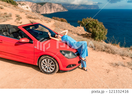 A woman is laying on top of a red convertible car. The car is parked on a dirt road near the ocean. The woman is wearing a blue dress and she is enjoying the moment. 121332416